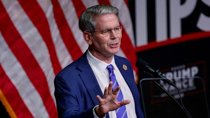 Scott Bessent speaks at a Republican party campaign event in August 2024. He is seen gesturing while standing at a podium, with an American flag in the background.